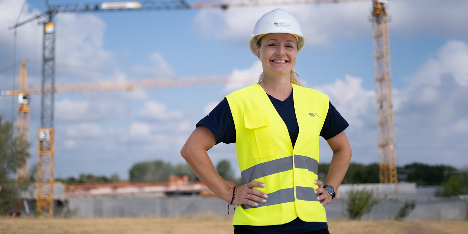 Eine Ingenieurin in neonfarbener Weste und Bauhelm steht stolz vor einer Baustelle