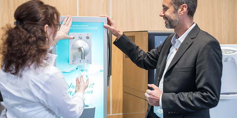 Ein Mitarbeiter der Berliner Wasserbetriebe führt einer Kundin einen Wasserspender vor